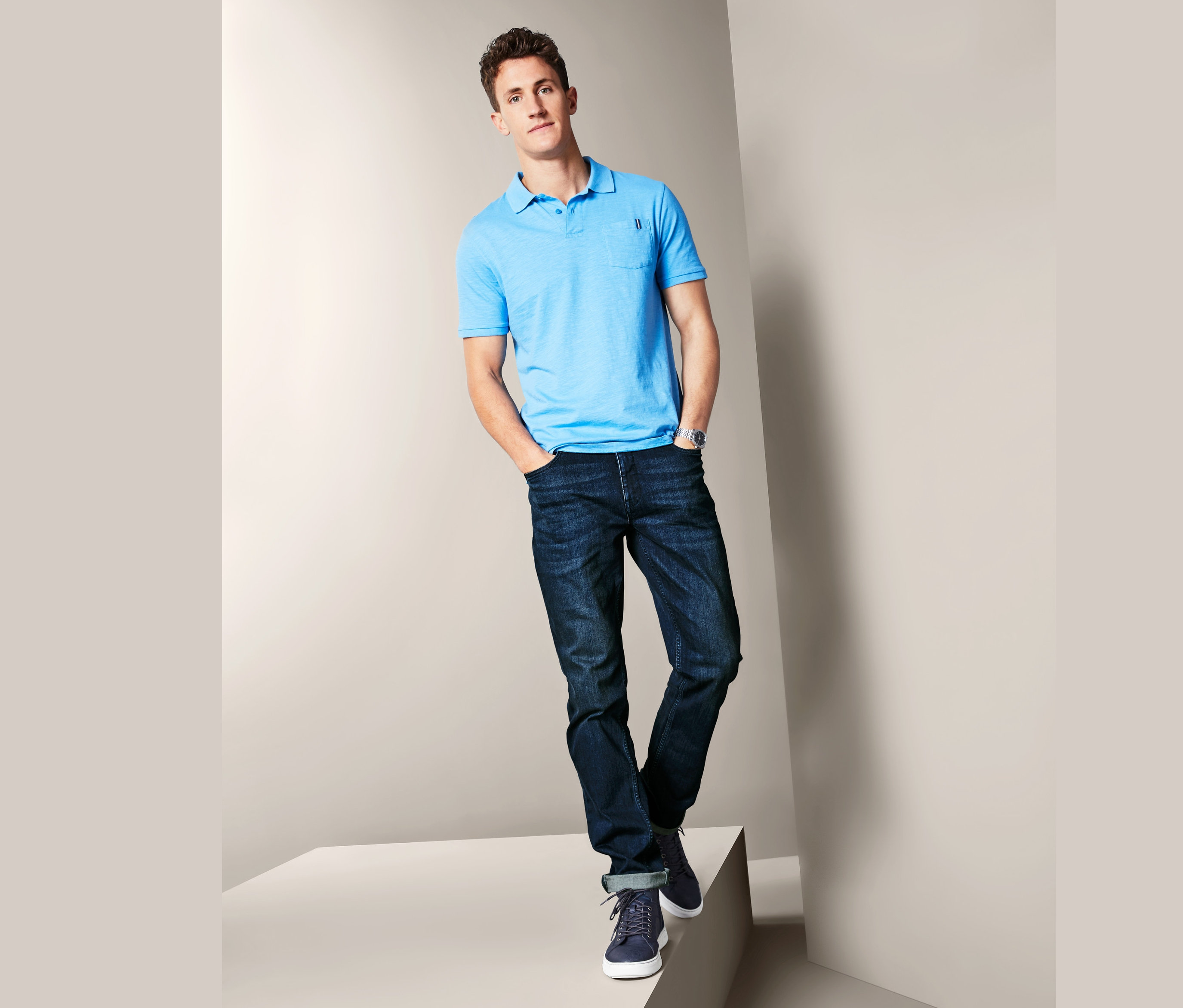 Mann posiert auf einem Podest und trägt Jeans und ein blaues Poloshirt.