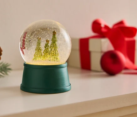 Schneekugel mit drei Bäumen auf einem grünen Sockel neben einem Tannenzapfen und einem Geschenk mit rotem Band.