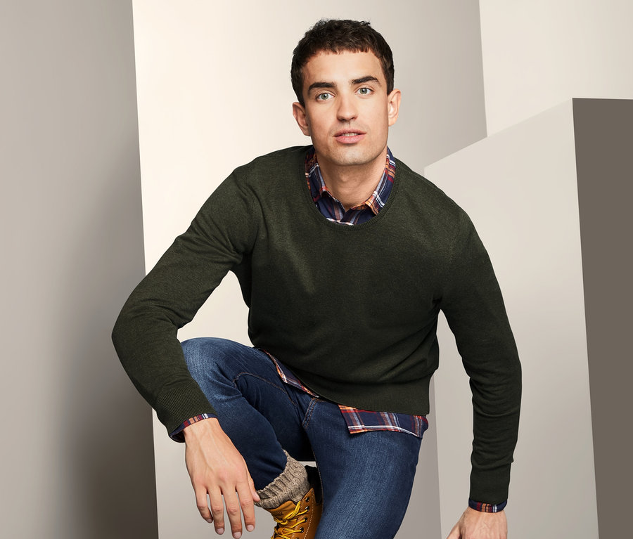 Mann posiert kniend und trägt einen Pullover mit Rundhalsausschnitt, Jeans und Stiefel.