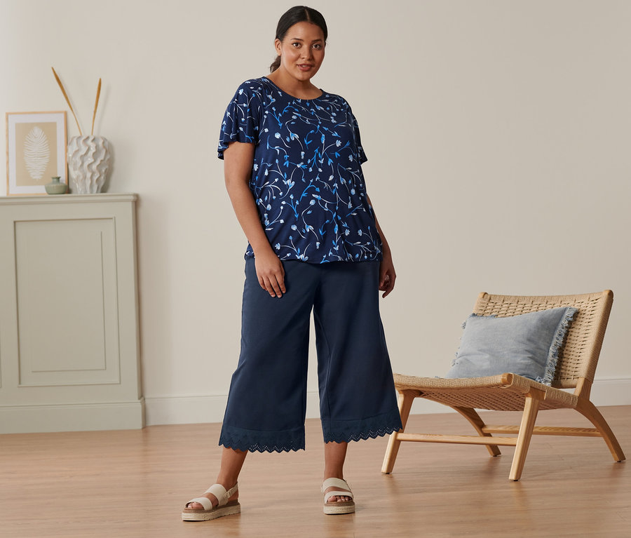 Eine Frau steht und trägt ein dunkelblaues Blusenshirt mit Flügelärmeln, eine Culotte mit Stickerei und Plateau-Sandalen aus Leder.