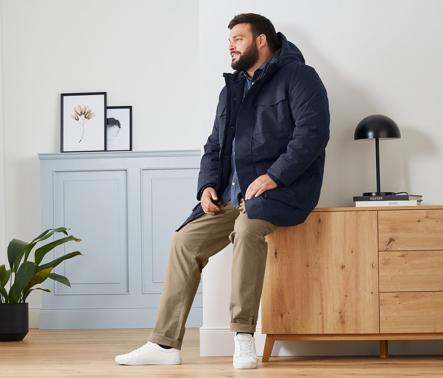 Ein Mann sitzt auf einer Holzkommode und trägt eine blaue Parka, ein Hemd Comfort Fit und eine camelfarbene Chino.