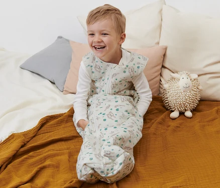 Lächelnder Junge sitzt in Schlafsack 2,5 TOG, 110 cm, mit Otter-Print auf dem Bett.