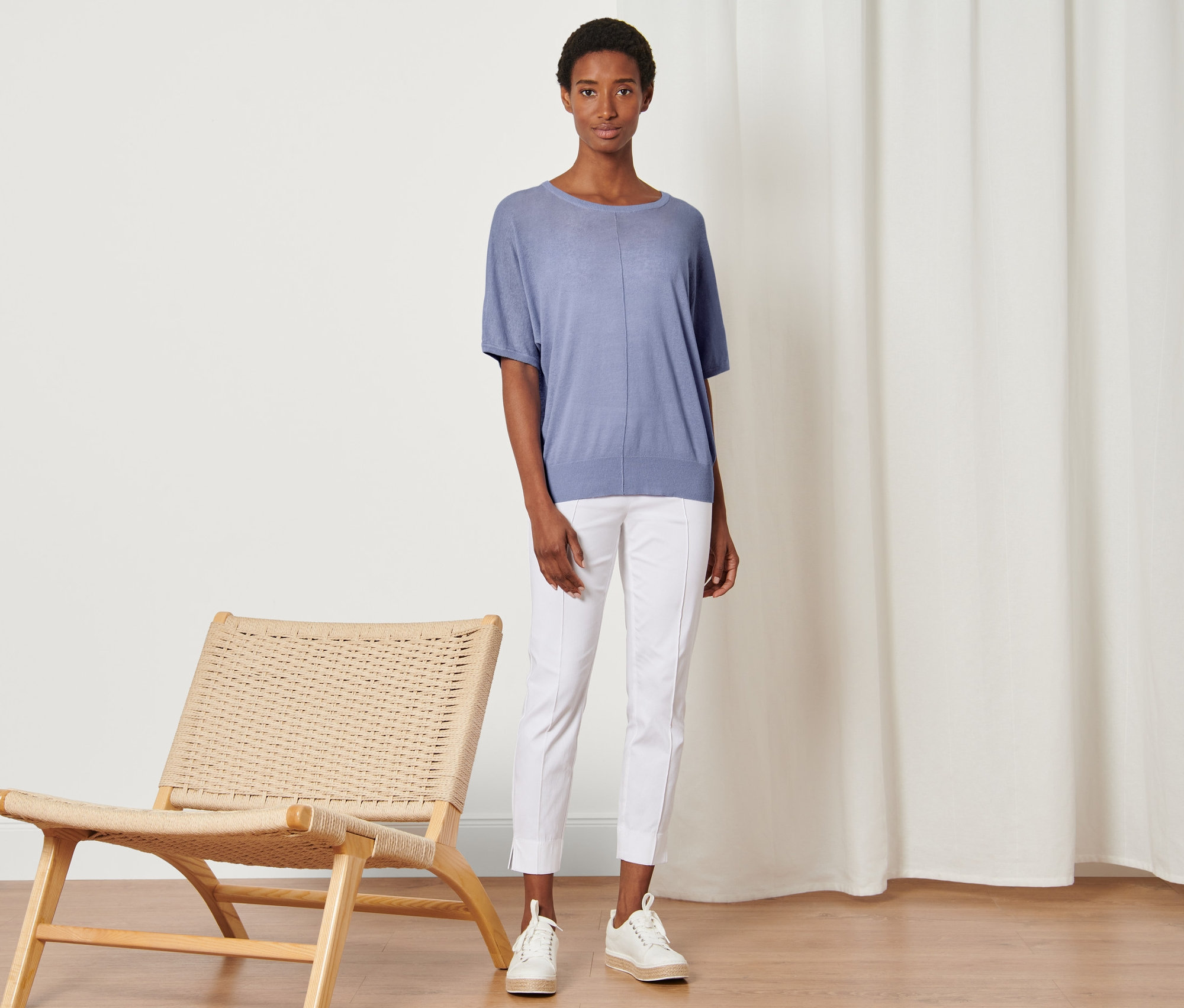 Frau steht neben einem Korbsessel und trägt ein Feinstrick-Shirt aus Leinenmix, weiße Hosen und Turnschuhe.
