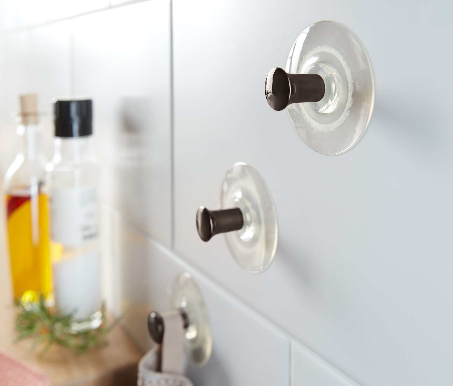 Drei mit Saugnapf befestigte Haken an weißen Fliesen, an einem Haken hängt ein graues Handtuch.