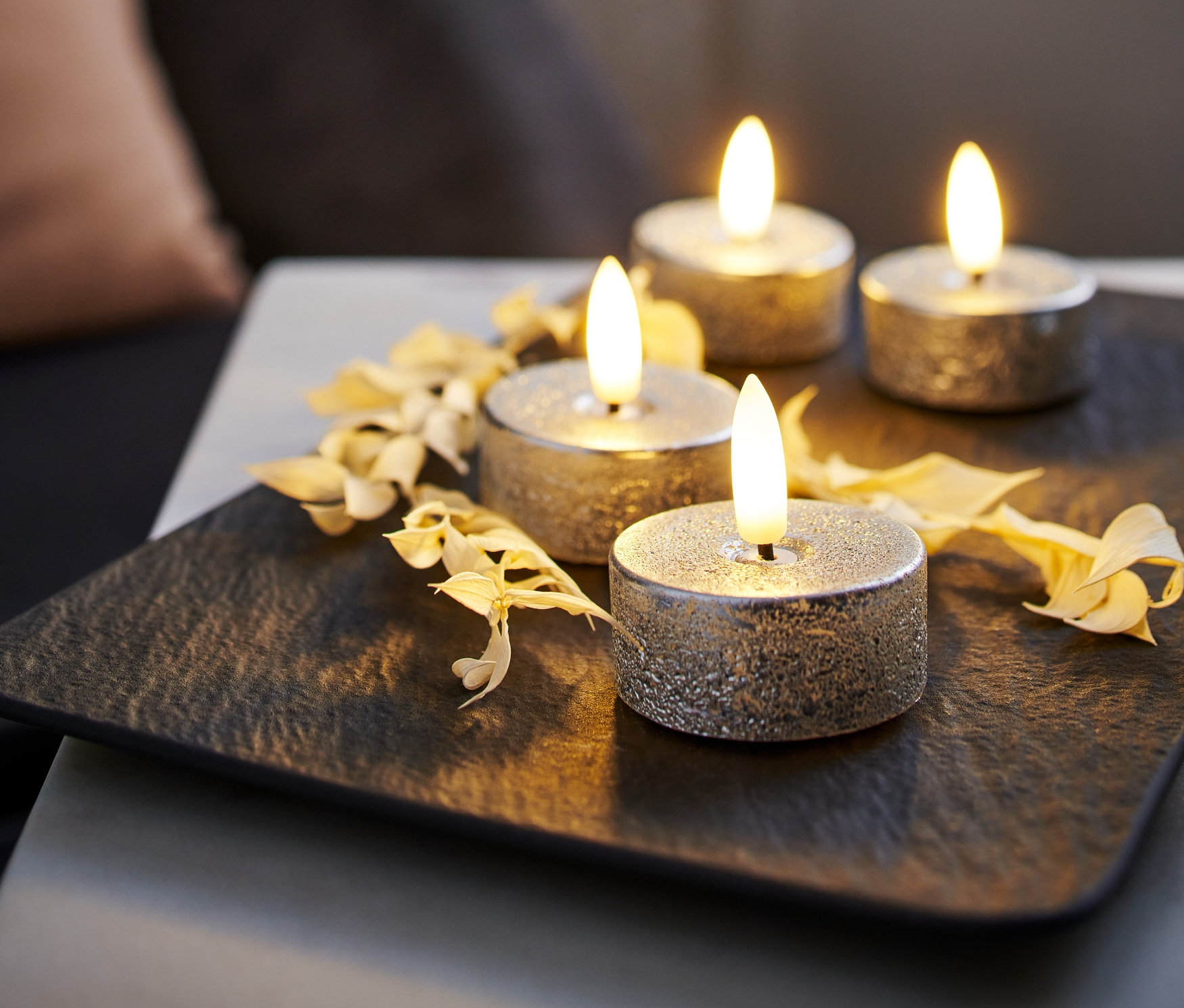 Vier silberfarbene LED-Echtwachs-Teelichter auf einer schwarzen Unterlage, dekoriert mit Trockenblumen.