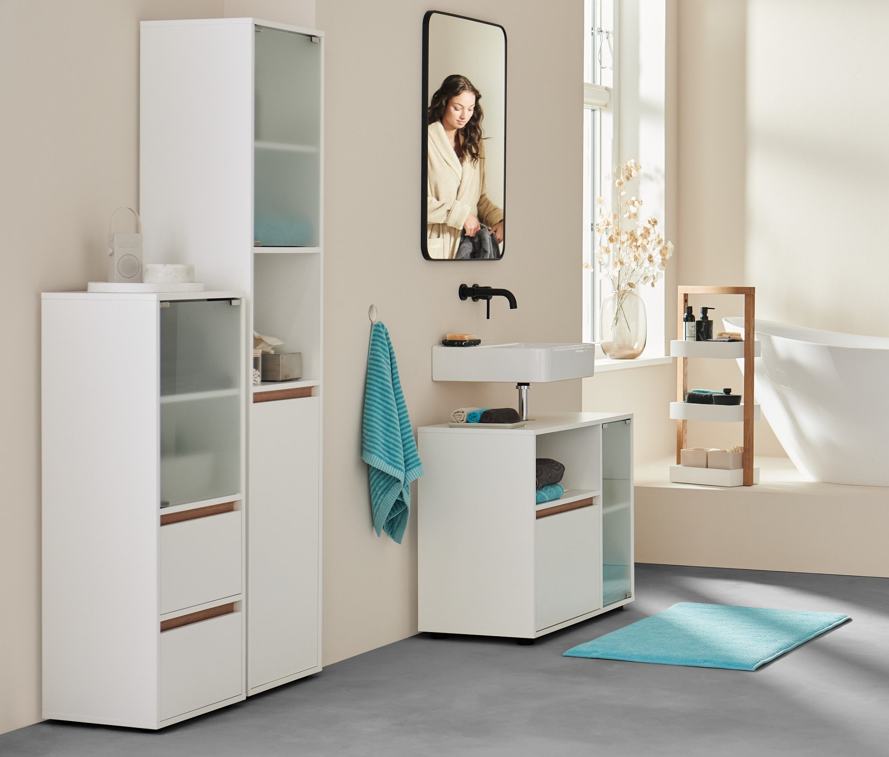 Im Badezimmer steht eine Frau im Bademantel an der Badewanne. Im Vordergrund Sideboard, Bad, extragroßes Badetuch in Anthrazit und zwei aqua Handtücher.