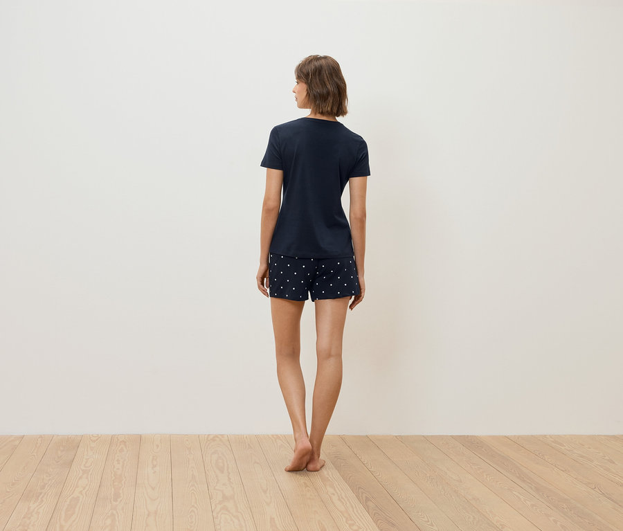 Eine Frau steht mit dem Rücken zur Kamera und trägt ein dunkelblaues T-Shirt und Shorts mit weißen Punkten.