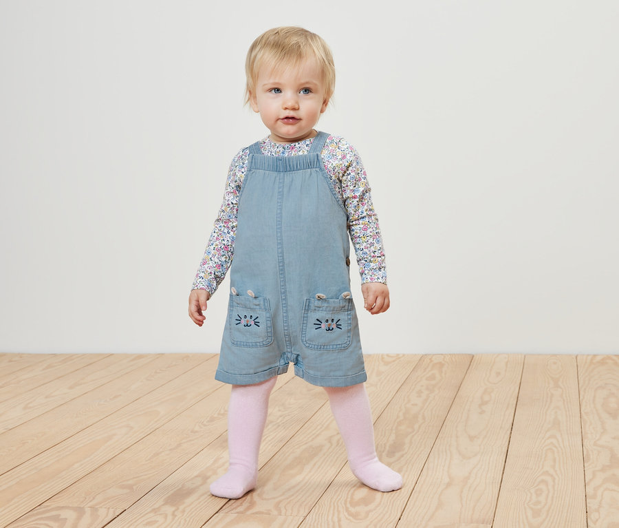 Kleinkind in Kinder Chambray-Latzhose und geblümten Oberteil steht auf Holzboden.