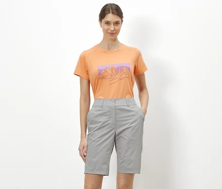 Eine Frau steht vor einem weißen Hintergrund und trägt ein orangefarbenes T-Shirt mit einer Abbildung von Bergen und graue Rad-und-Wandershorts »Pro«.