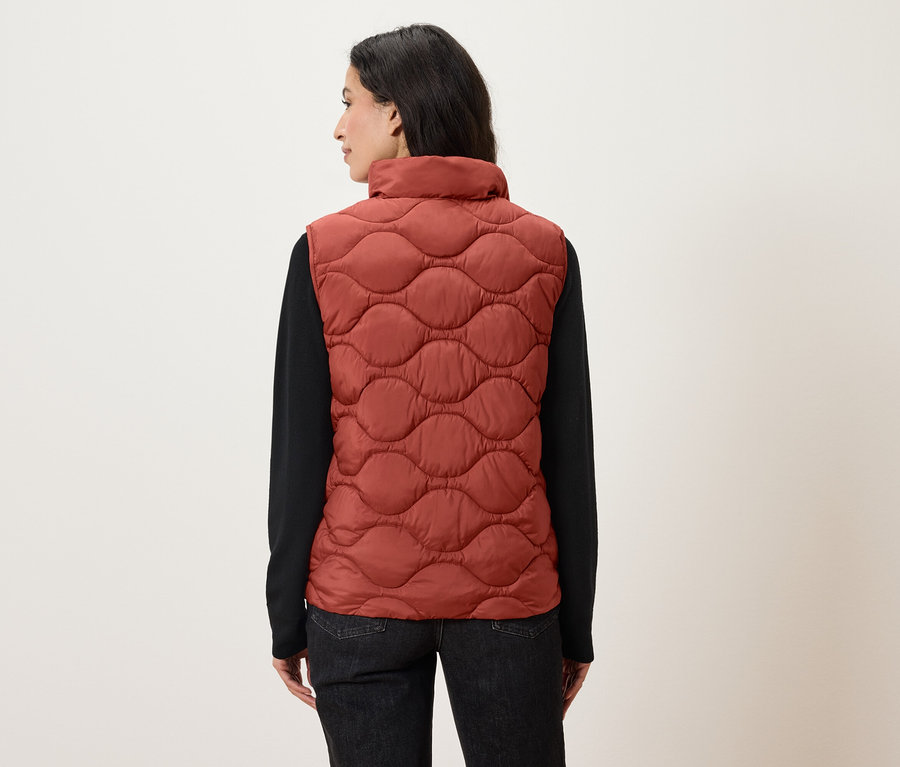 Frau mit dem Rücken zur Kamera trägt eine orangefarbene Steppweste und ein schwarzes Langarmshirt.