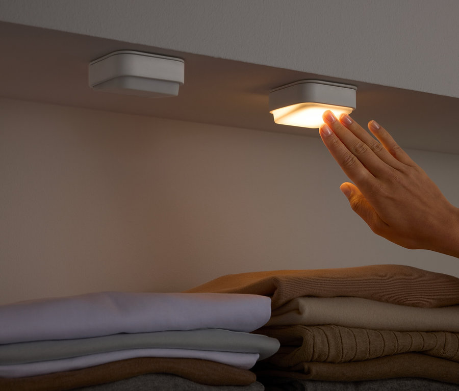Eine Hand berührt ein quadratisches Licht unter einem Regal über gefalteter Kleidung.