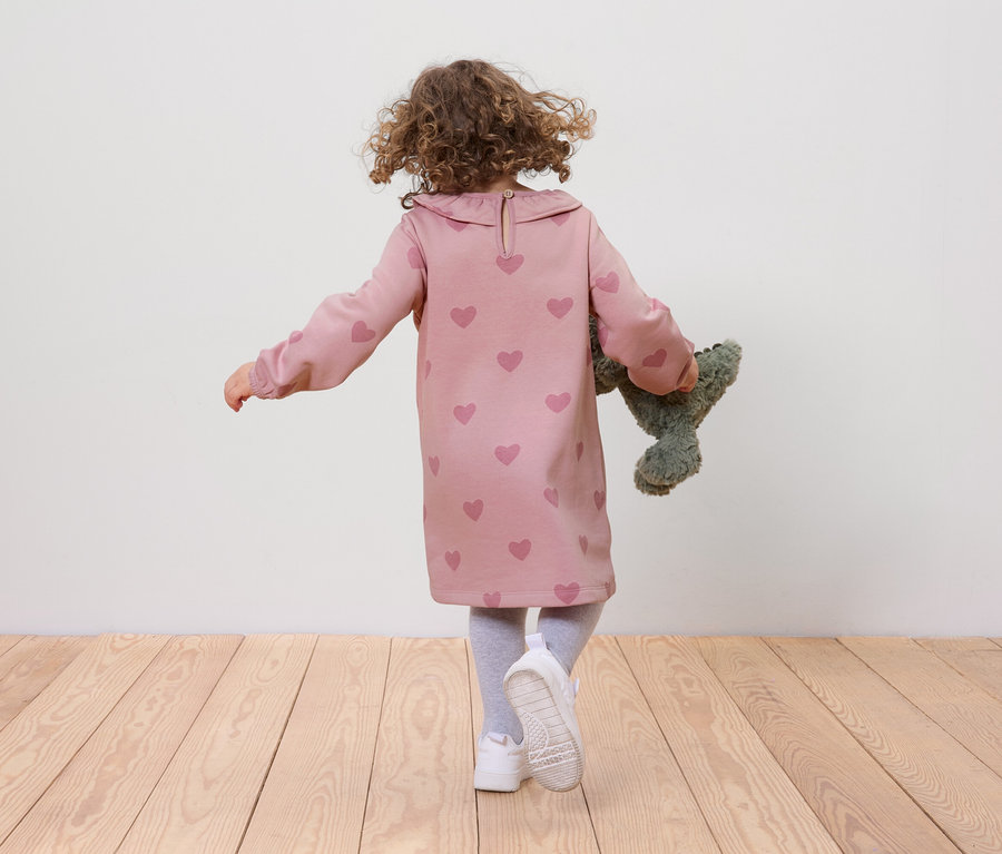 Ein Mädchen mit lockigem Haar, bekleidet mit einem rosa Kleid mit Herzen und weißen Schuhen, geht auf einem Holzboden und hält einen grünen Teddybären.