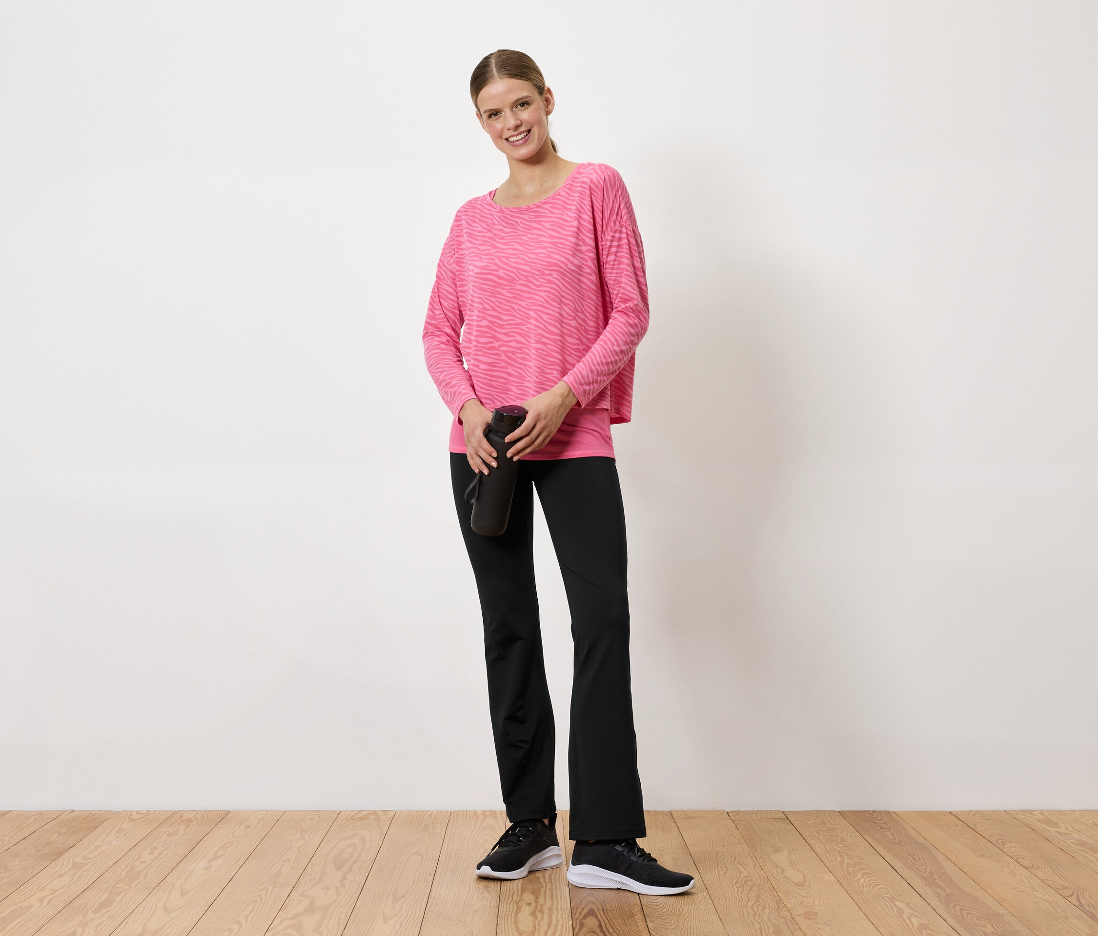 Frau in pinkem Pullover und schwarzen Hosen hält eine schwarze Wasserflasche in den Händen.