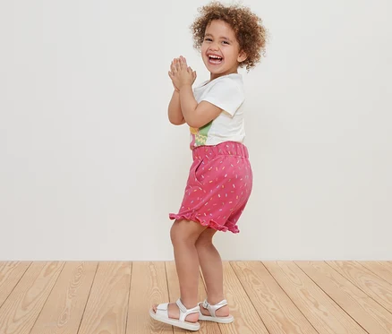 Lächelndes lockiges Mädchen klatscht in die Hände und trägt Kinder-Sweat-Shorts.