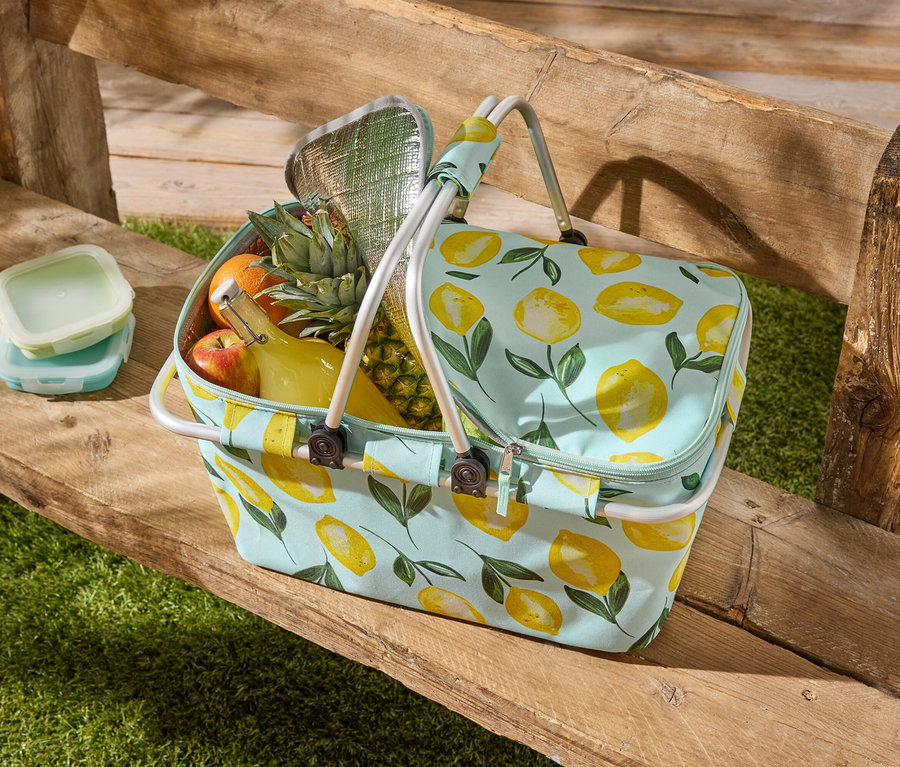 Eine Picknicktasche mit Zitronenmuster steht auf einer Holzbank, gefüllt mit Obst, einer Flasche und zwei Frischhaltedosen.
