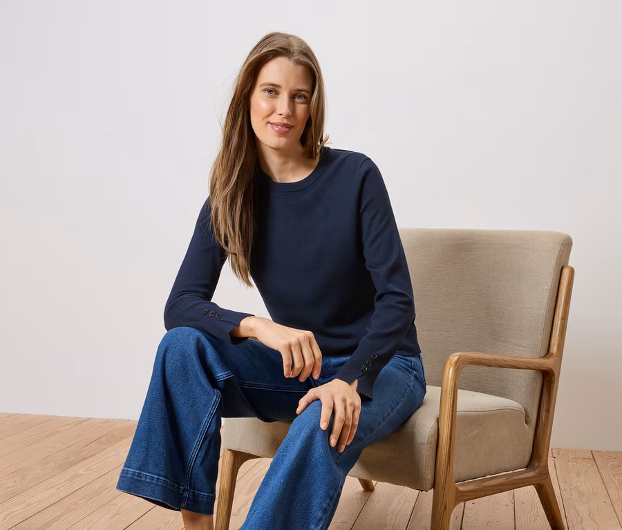 Frau mit langem Haar sitzt auf einem Stuhl und trägt ein marineblaues Langarmshirt und eine weite Jeans.