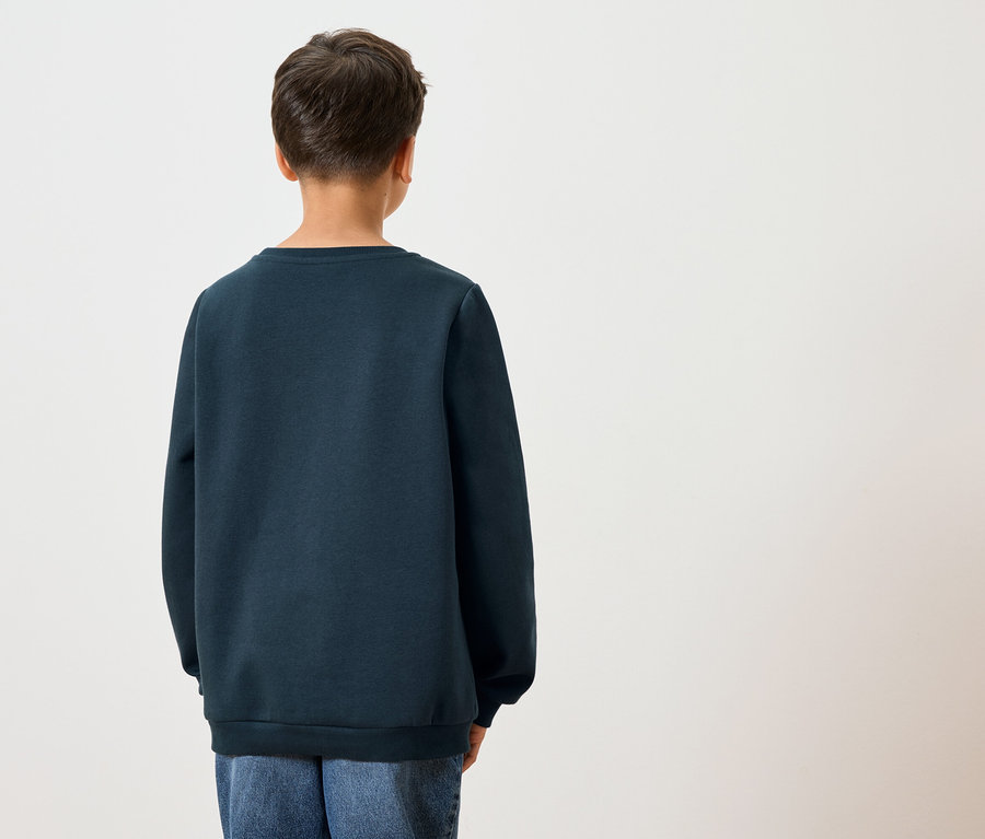 Rückansicht eines Kindes mit dunklen Haaren, das einen dunkelblauen Pullover und blaue Jeans vor einem weißen Hintergrund trägt.