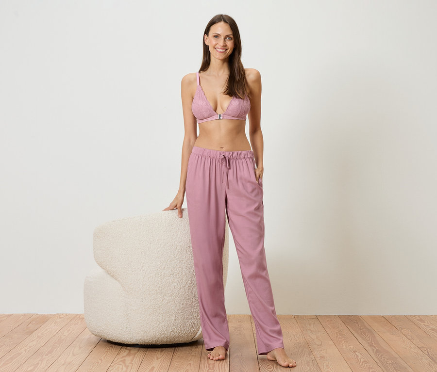 Frau steht barfuß neben einem weißen Sessel, bekleidet mit einem rosa Spitzen-BH und einer locker sitzenden rosa Hose.