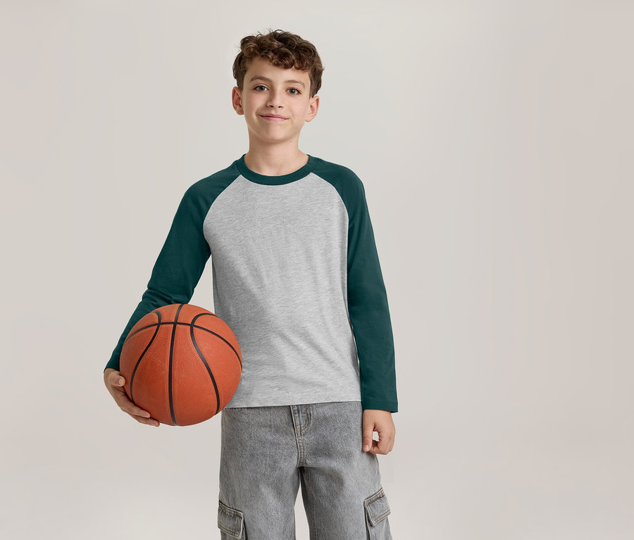 Ein lächelnder Junge in einem grau-dunkelgrünen Langarmshirt und grauen Cargohosen hält einen Basketball in seiner linken Hand.