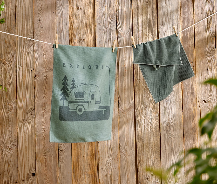 Ein grünes Geschirrtuch mit Wohnwagen-Motiv hängt neben einem strukturierten Handtuch an einer Wäscheleine vor einer Holzwand.