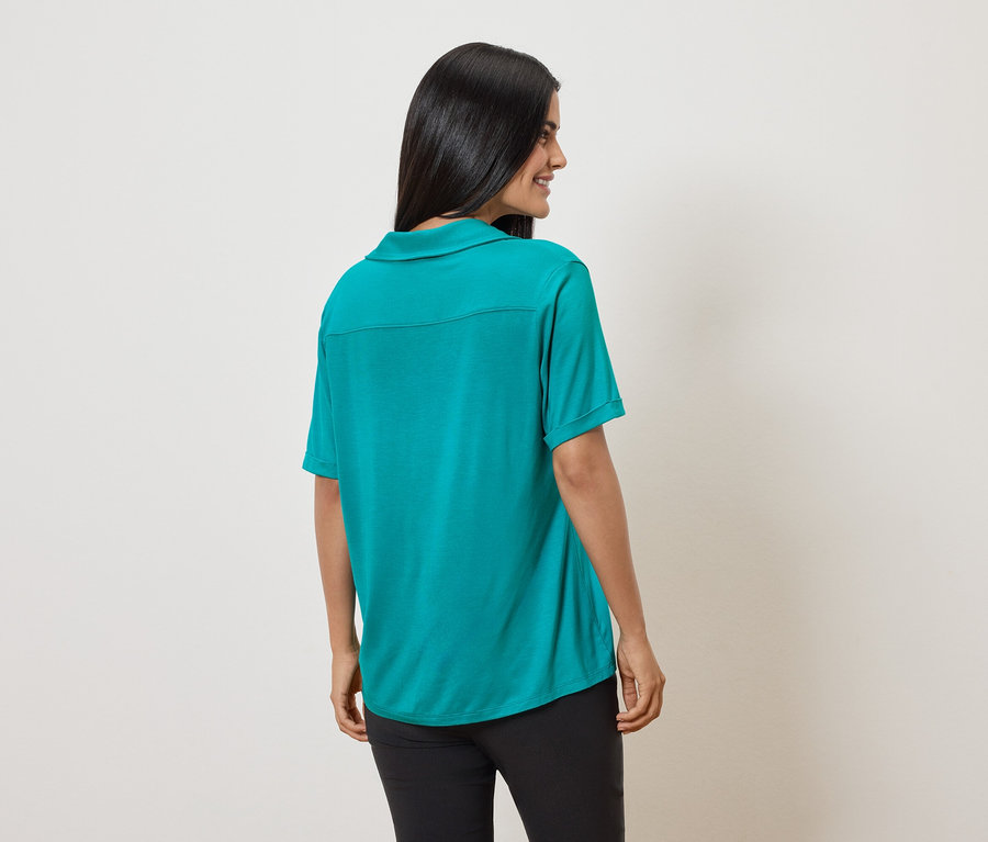 Rückansicht einer Frau mit dunklen Haaren, die ein grünes Poloshirt und schwarze Hosen vor einem cremefarbenen Hintergrund trägt.