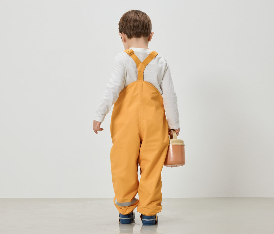 Ein Kind in gelber Latzhose und weißem Langarmshirt steht mit dem Rücken zur Kamera und hält eine Dose.