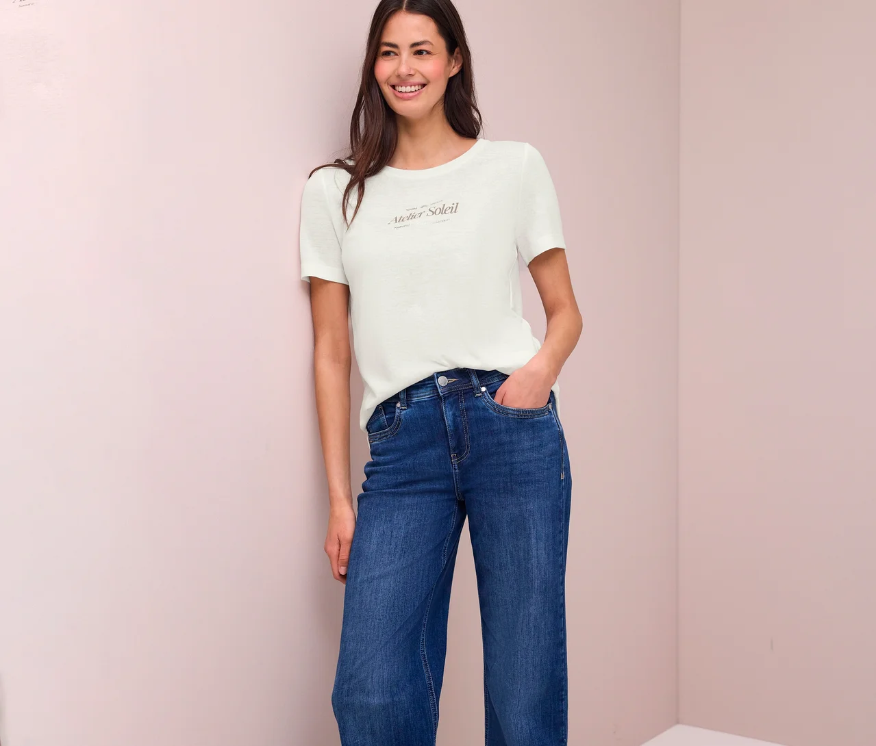 Lächelnde Frau mit Jeans und T-Shirt mit Aufdruck "Atelier Soleil" lehnt an einer Wand.