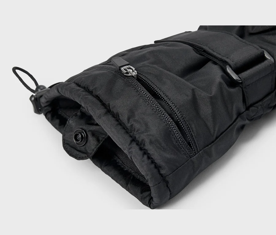 Nahaufnahme eines schwarzen, gepolsterten Handschuhs oder Ärmels mit teilweise geöffnetem Reißverschluss, einem Kordelzug mit Stopper, einem Druckknopf und einem verstellbaren Riemen mit Schnalle.