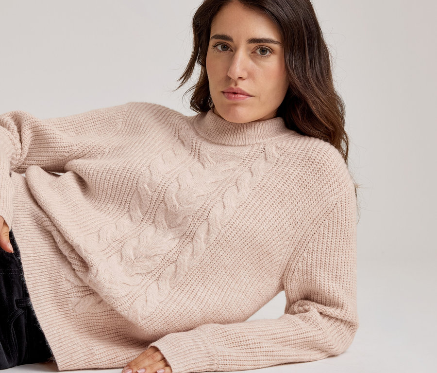 Liegende Frau in hellrosa Zopfstrickpullover mit Stehkragen und dunkler Hose.
