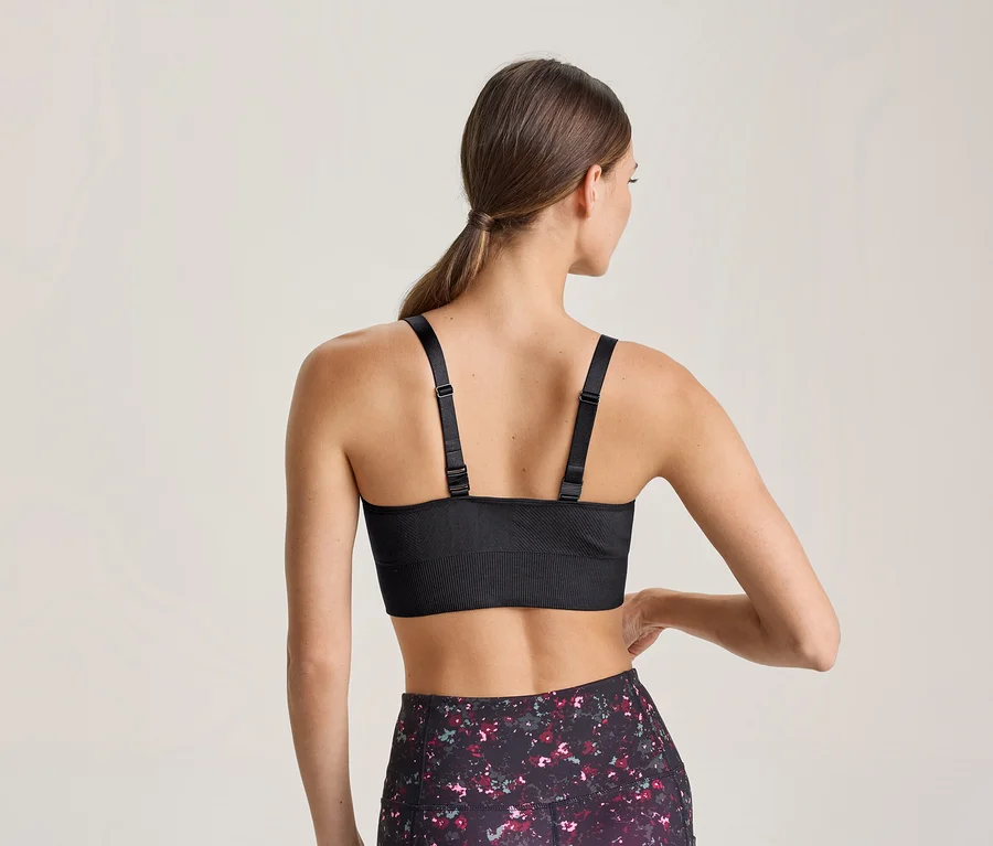 Frau von hinten, die einen schwarzen Sport-BH mit verstellbaren Trägern und gemusterte Leggings trägt. Ihre linke Hand ruht auf ihrer Hüfte.