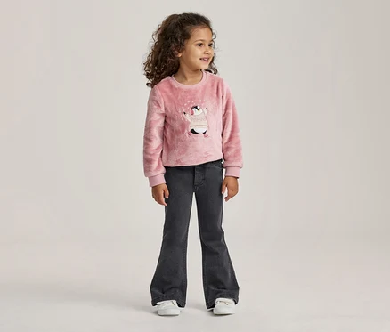 Junges Mädchen mit lockigem Haar steht da und trägt einen rosa flauschigen Pullover mit Pinguin-Motiv und dunkelgraue Schlagjeans.
