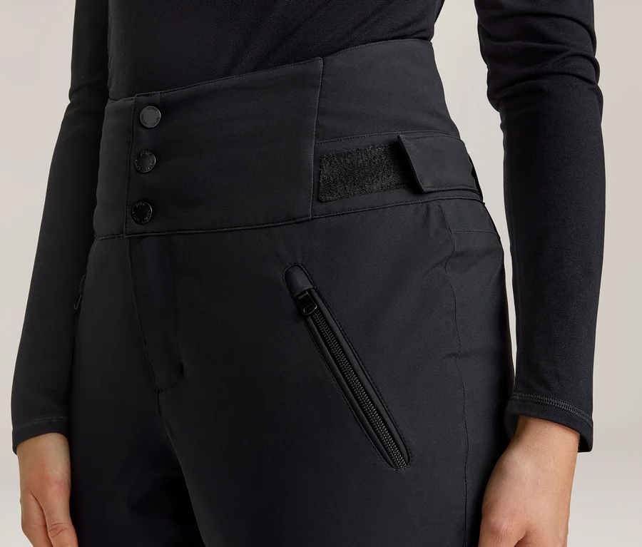 Nahaufnahme einer Person in einer schwarzen, hoch taillierten Hose mit drei Knöpfen und einem Klettverschluss am Bund sowie einer Reißverschlusstasche am rechten Oberschenkel. Dazu trägt die Person ein schwarzes Langarmshirt.