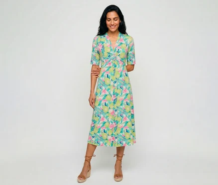 Frau mit dunklem Haar lächelt, trägt ein Olsen Jerseykleid mit buntem Zitronen- und Blumenmuster, V-Ausschnitt und gedrehter Taillendetail, dazu braune Keilsandalen.