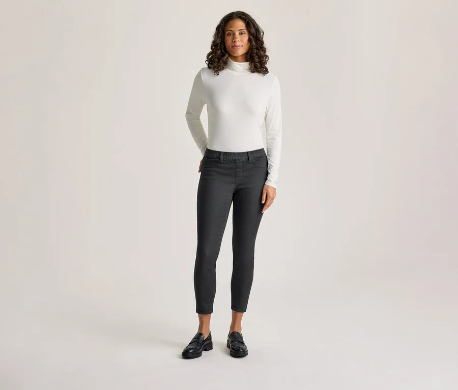 Eine Frau mit dunklem, lockigem Haar steht da und trägt ein weißes, langärmeliges Rollkragenshirt und dunkelgraue Treggings - Fit »Nelly«, mit den Händen in den Taschen.