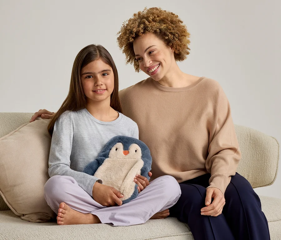 Eine erwachsene Frau und ein junges Mädchen sitzen lächelnd auf einem Sofa. Das Mädchen links hält ein blau-beiges Pinguin-Stofftier. Die Frau rechts hat ihren Arm um das Mädchen gelegt.