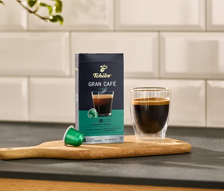 Auf einem Holzbrett stehen eine Packung Gran Café Lungo, eine grüne Kapsel und ein Glas Kaffee.