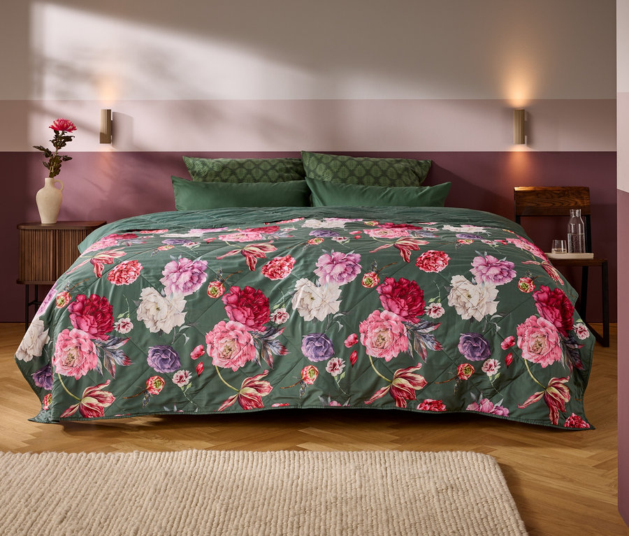 Ein Bett mit einer dunkelgrünen, gesteppten Tagesdecke mit großen rosa, roten und weißen Blumen. Vier grüne Kissen liegen am Kopfende, flankiert von Holznachttischen und einer zweifarbigen Wand.