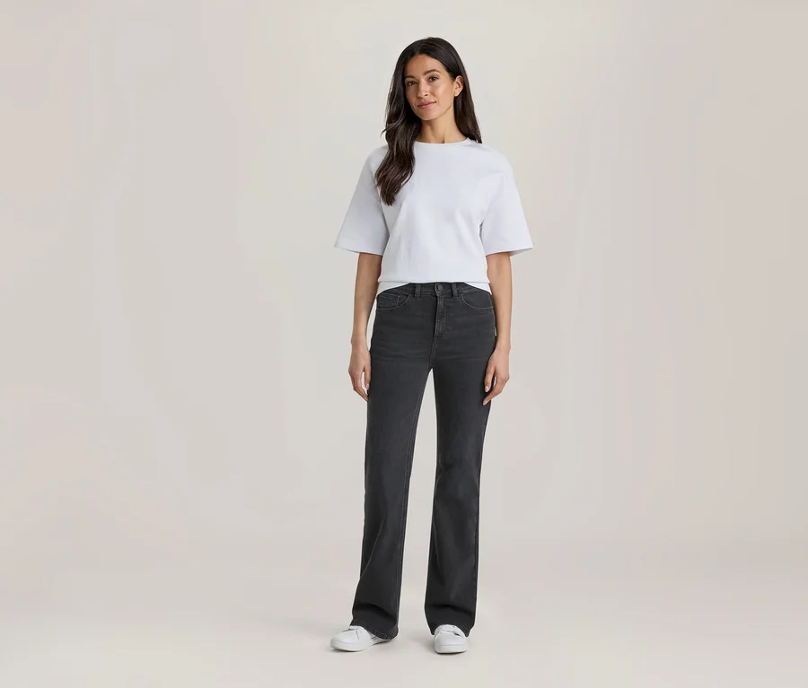 Frau steht in einem weißen T-Shirt, dunkelgrauen Jeans und weißen Turnschuhen.