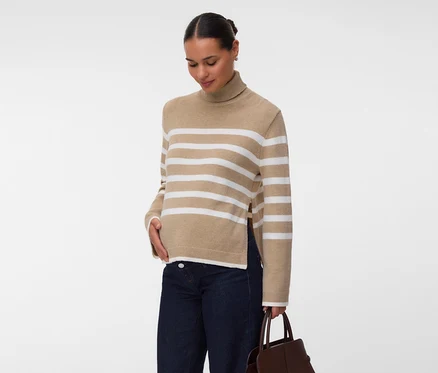 Frau in einem beigefarbenen Rollkragenpullover mit weißen Streifen und Seitenschlitzen, dunklen Jeans und einer braunen Handtasche, die Hand auf dem Bauch.