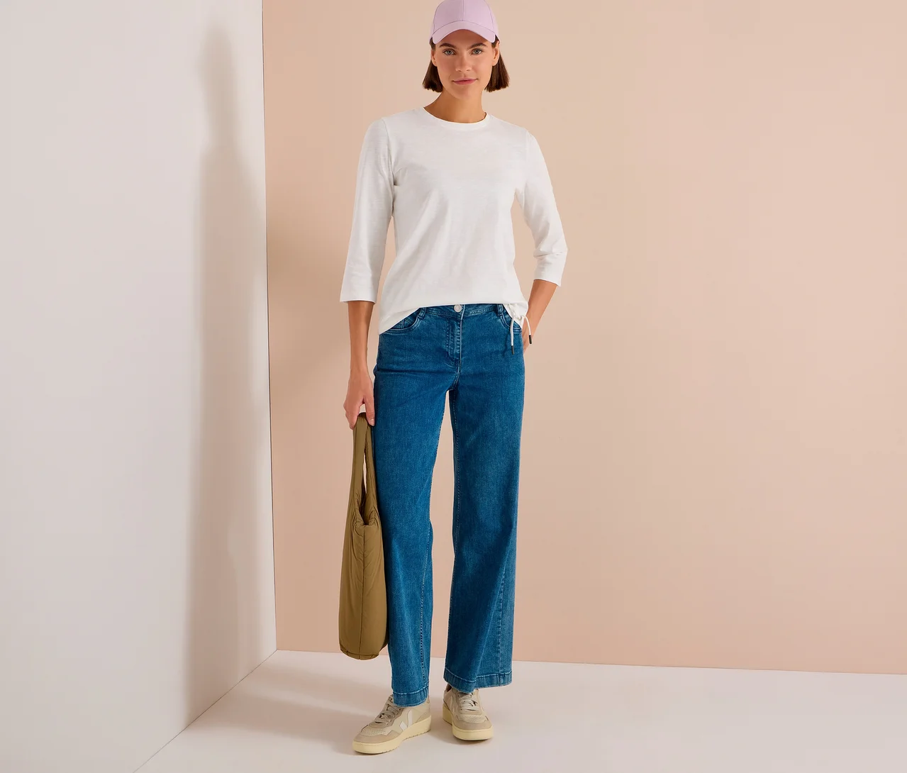 Frau trägt ein weißes CECIL 3/4- Shirt, blaue Jeans, eine hellviolette Kappe, helle Sneaker und hält eine grüne Tragetasche.