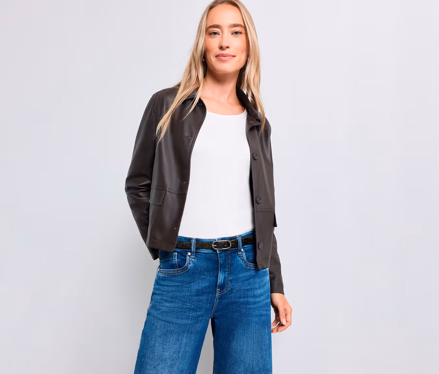 Eine blonde Frau steht lächelnd vor einem weißen Hintergrund. Sie trägt eine dunkelbraune Jacke, ein weißes Oberteil, blaue Jeans und einen dunklen Gürtel.