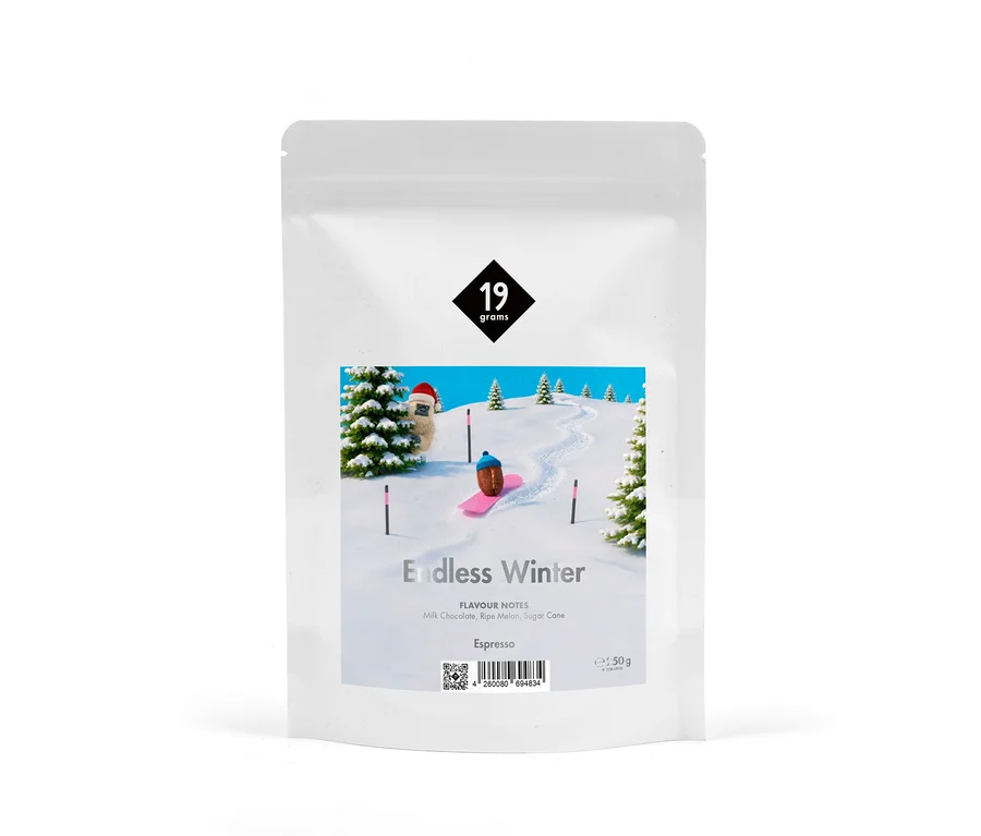 Weißer Kaffeebeutel mit Etikett, das eine Winterlandschaft zeigt: eine Kaffeebohne auf einem pinken Snowboard, eine pelzige Figur mit Weihnachtsmütze und Tannenbäume auf verschneiten Hügeln. Text: „19 grams“, „Endless Winter“, „Espresso“ und Geschmacksnoten.