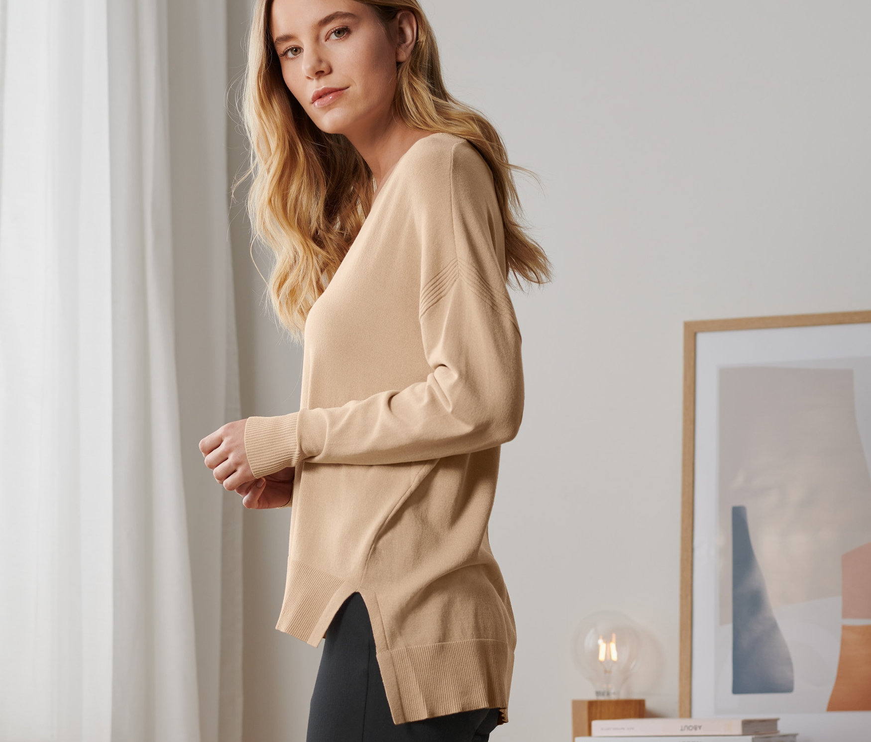 Blonde Frau posiert in beigem Feinstrick-Pullover und schwarzen Hosen.