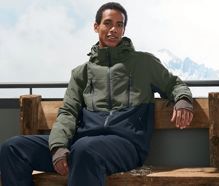 Mann sitzt auf einer Bank und trägt eine khakifarbene Skijacke und Skischuhe.