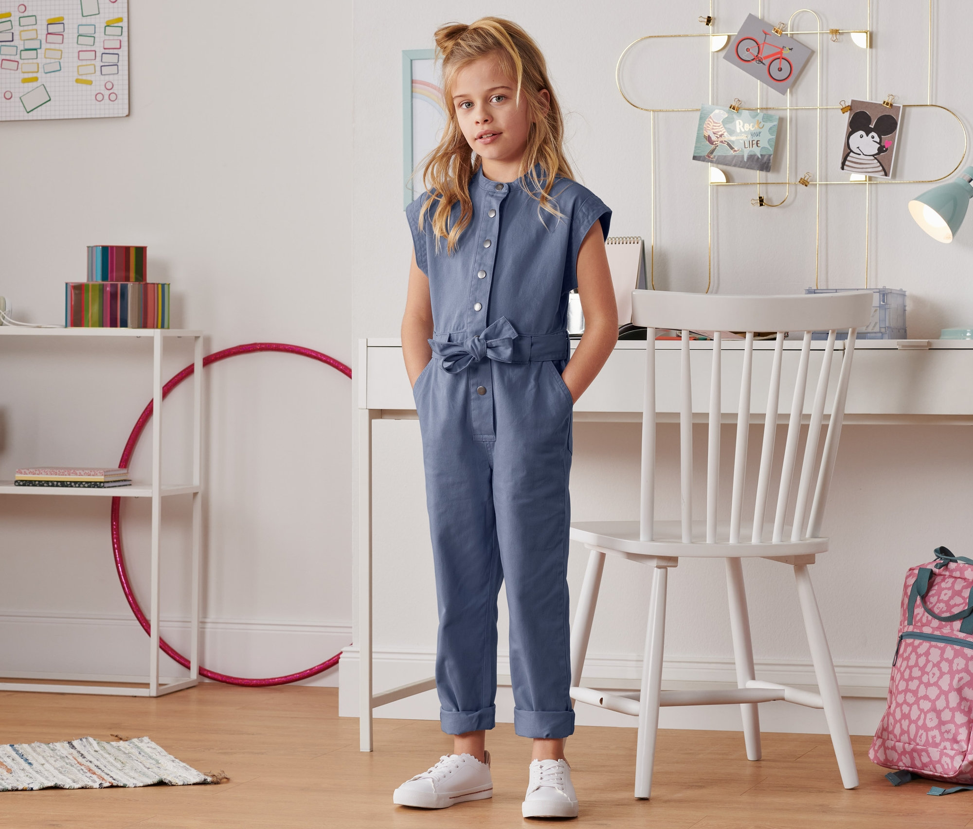 Mädchen steht in blauem Overall in einem Kinderzimmer.