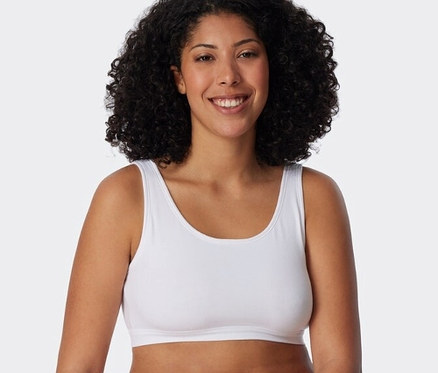 Lächelnde Frau mit lockigem Haar trägt ein weißes SCHIESSER Bustier und weiße Slips, an einem weißen Block lehnend.