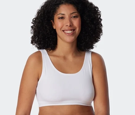 Lächelnde Frau mit lockigem Haar trägt ein weißes SCHIESSER Bustier und weiße Slips, an einem weißen Block lehnend.