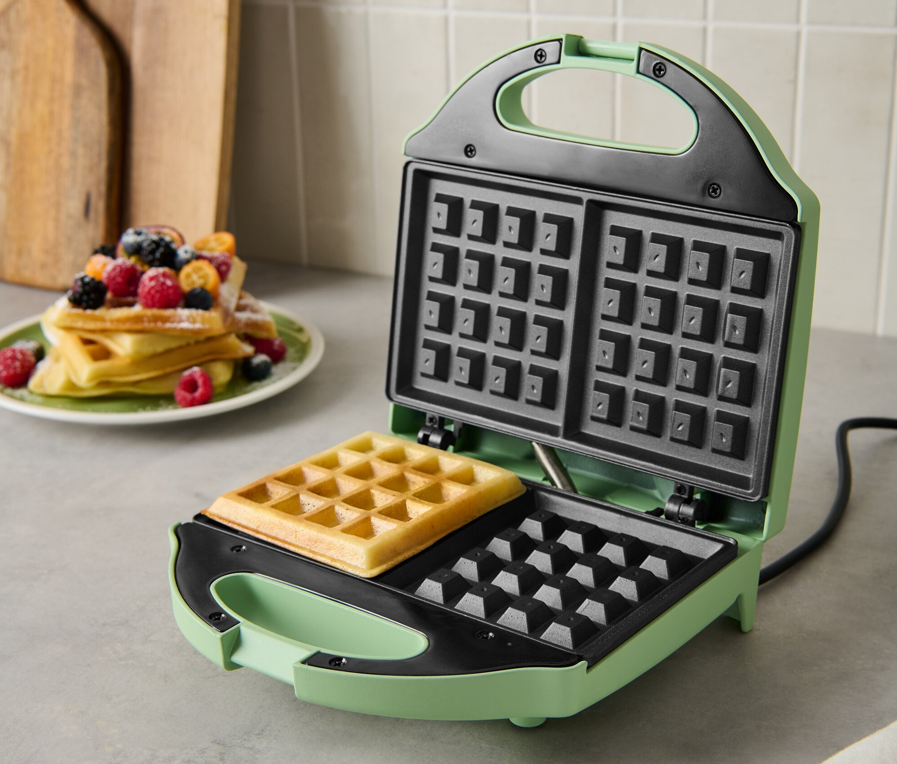 Ein grünes Waffeleisen mit einer gebackenen Waffel darin. Auf einem Teller liegt ein Stapel Waffeln mit Früchten.