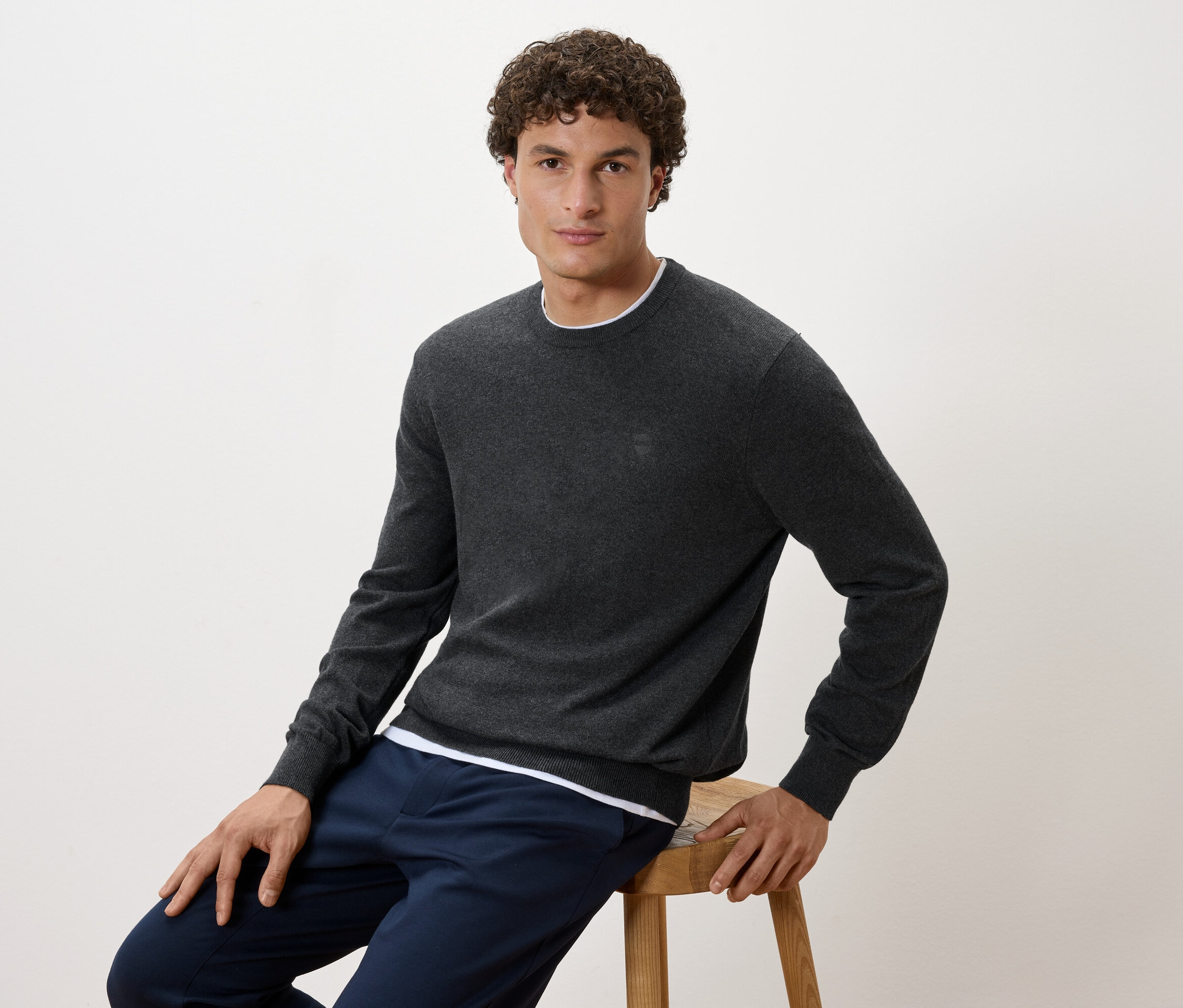 Mann sitzt auf einem Hocker, trägt einen grauen Pullover und dunkelblaue Hosen.