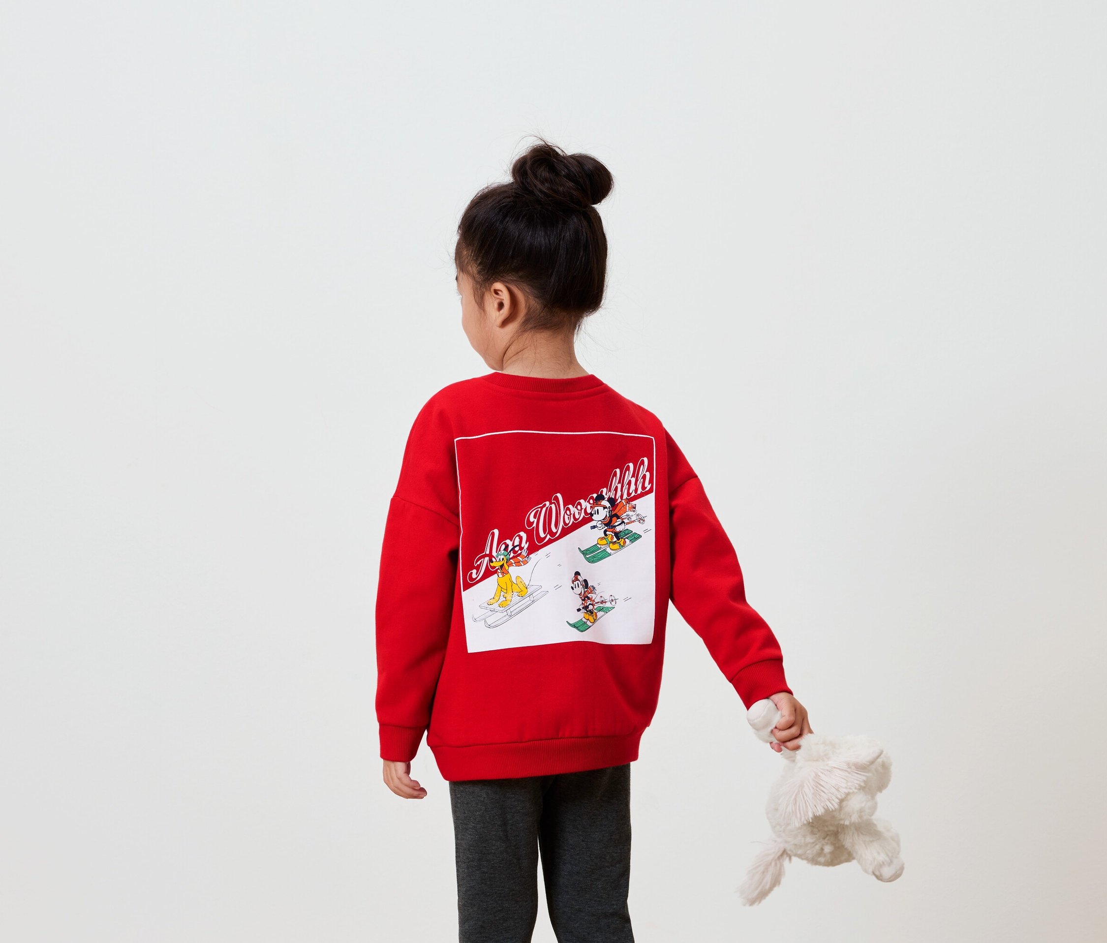 Mädchen mit dem Rücken zur Kamera trägt ein rotes Sweatshirt mit einem Bild auf dem Rücken und hält ein Plüschtier in der Hand.